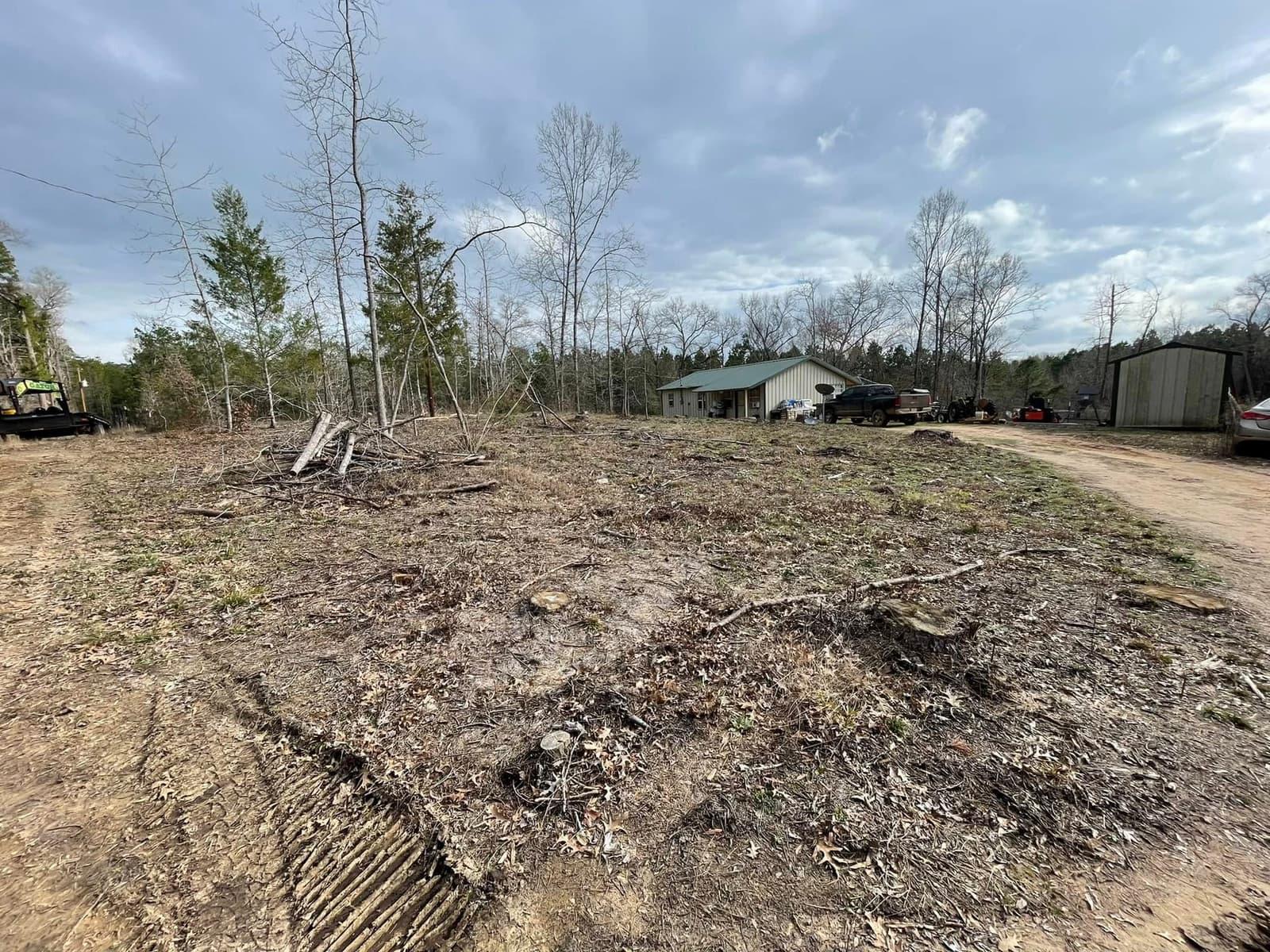 Homesite freshly cleared of brush next to a small house, debris staged and ground opened up before pad grading